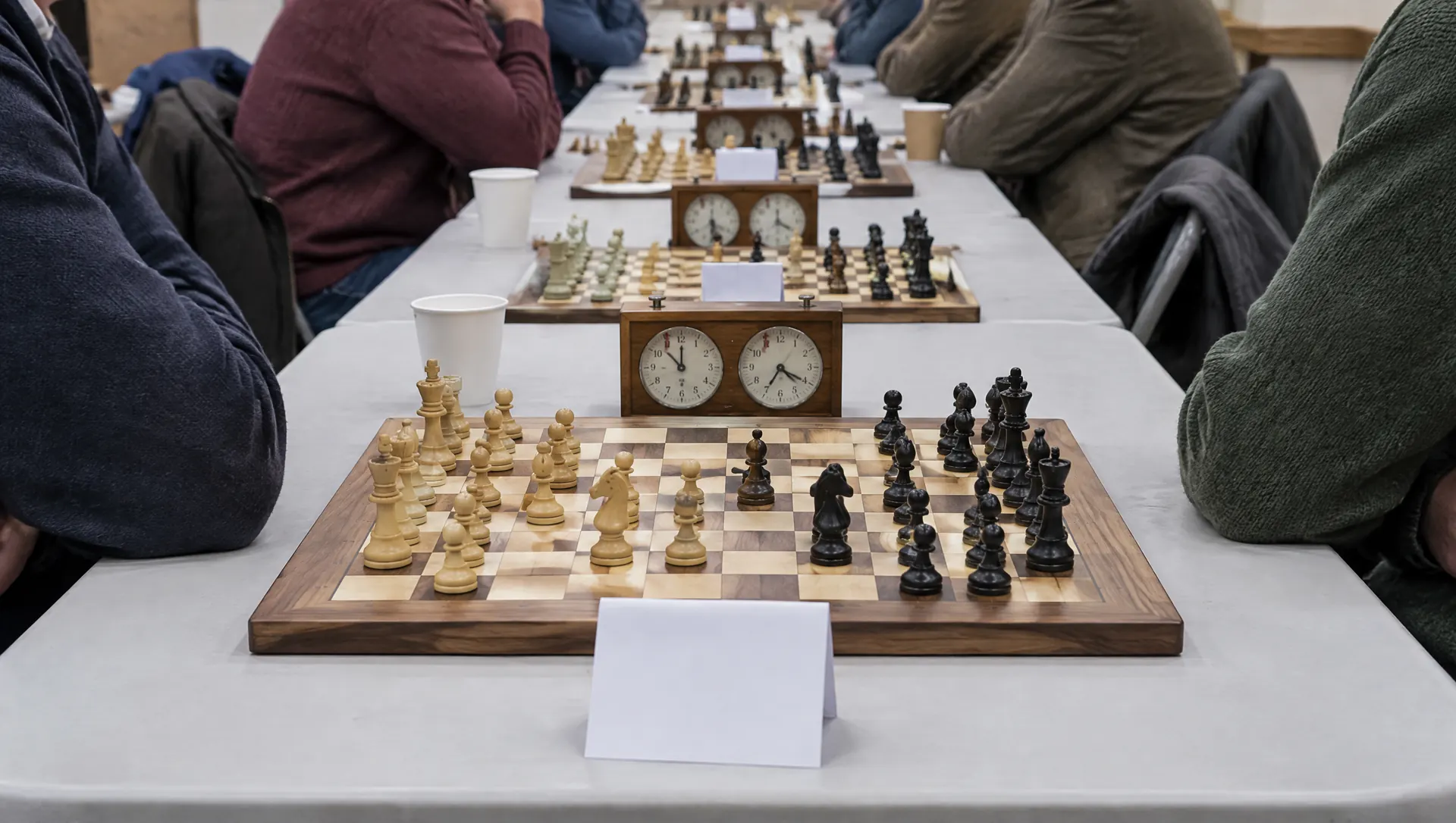 A long table at a chess club night: wooden sets in play, a chess clock beside every board and a blank table tent at the nearest, players on both sides seen only at the shoulders, paper cups and coats on folding chairs.
